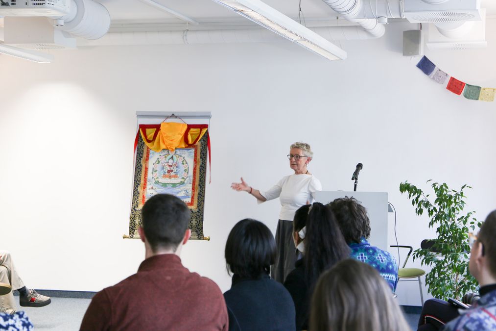 enlarge the image: Woman speaks in front of an audience and points to a colourful Buddhist scroll painting